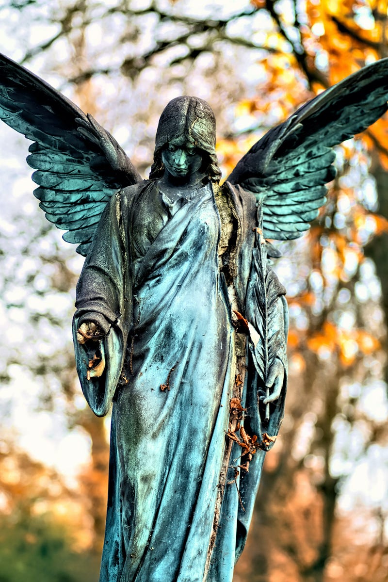 Statue of dramatic angel looking downward with trees in background.