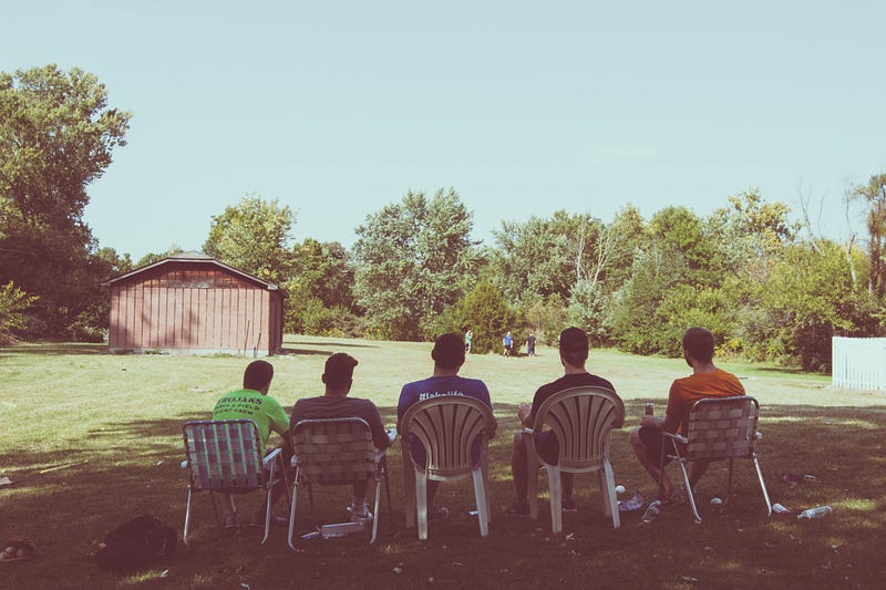 Men sitting on lawn chairs looking out at a field with guys in the background.