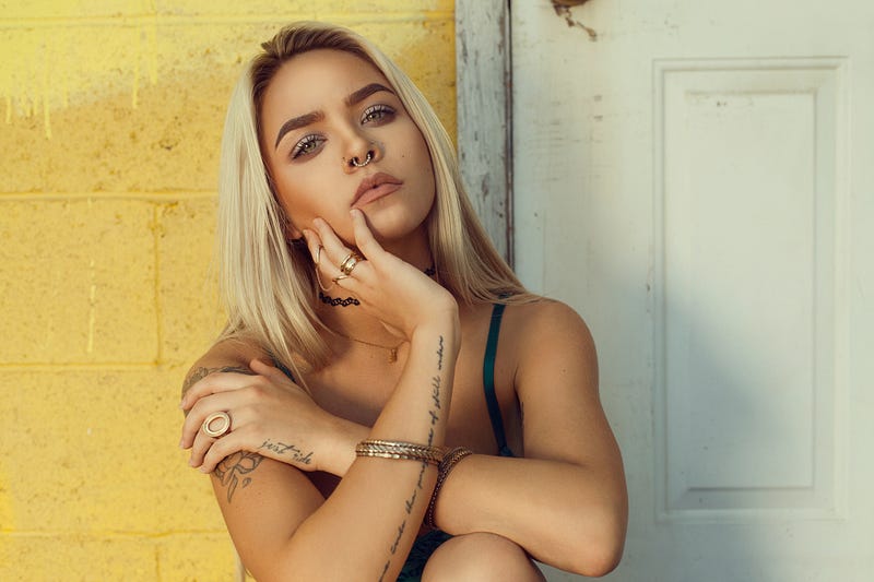 A young woman with blond hair, a nose ring and writing tattooed on her arms and hand, stares ahead pensively.